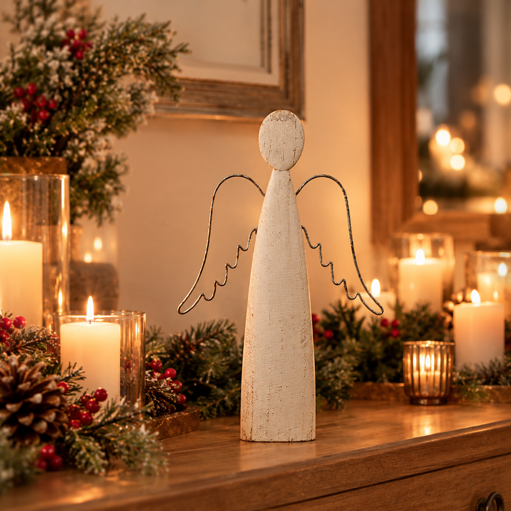 A rustic white wooden angel figurine with wire wings stands on a wooden table surrounded by lit candles, pinecones, and greenery in a warm holiday setting.