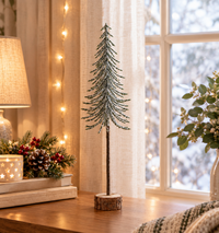 A decorative artificial Christmas tree with a textured pine-green body and a brown trunk on a round wood slice base, styled on a wooden table in a cozy holiday setting with warm string lights, a lamp, and a window view.