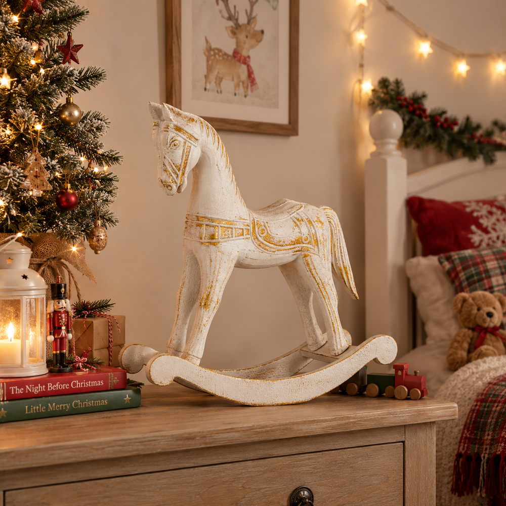 A distressed white rocking horse with gold decorative accents sits on a wooden bedside table in a cozy holiday bedroom. The scene includes a lit Christmas tree, string lights, framed art, and a teddy bear on pillows in the background.