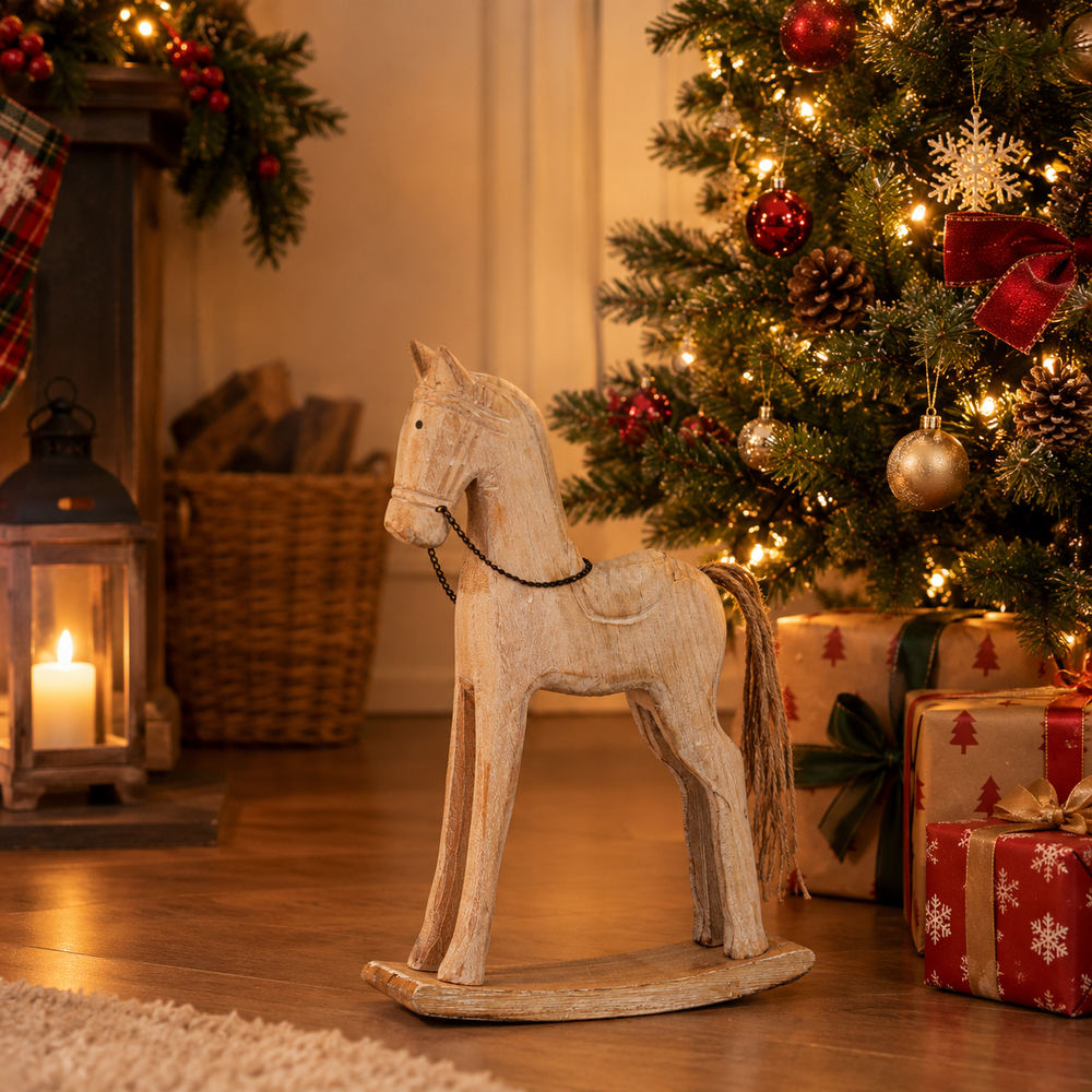A rustic wooden rocking horse with a weathered off-white finish stands on a wooden floor in a cozy holiday setting, positioned in front of a decorated Christmas tree with wrapped presents and a lit lantern.