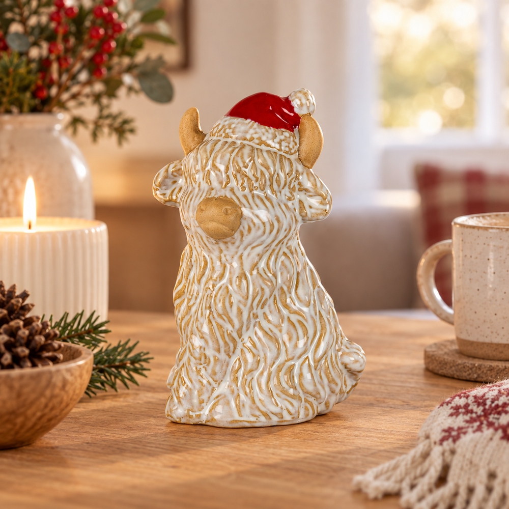 A ceramic Highland cow figurine with textured cream and gold fur, wearing a red Santa hat, displayed on a wooden table in a cozy holiday setting with a candle, pinecones, and a mug.
