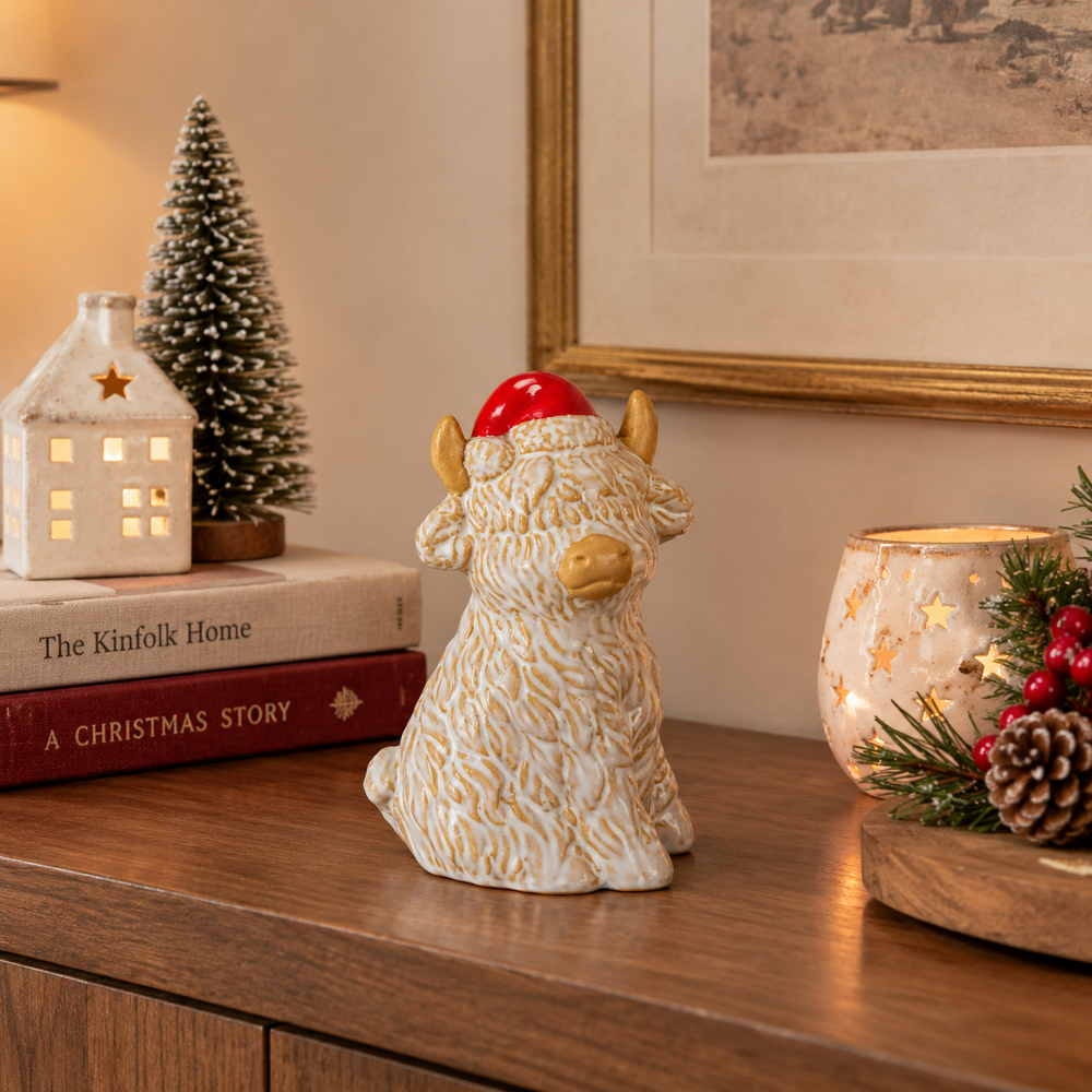 A ceramic Scottish Highland cow figurine with textured cream and gold detailing, wearing a red Santa hat, displayed on a wooden console table in a cozy holiday setting.