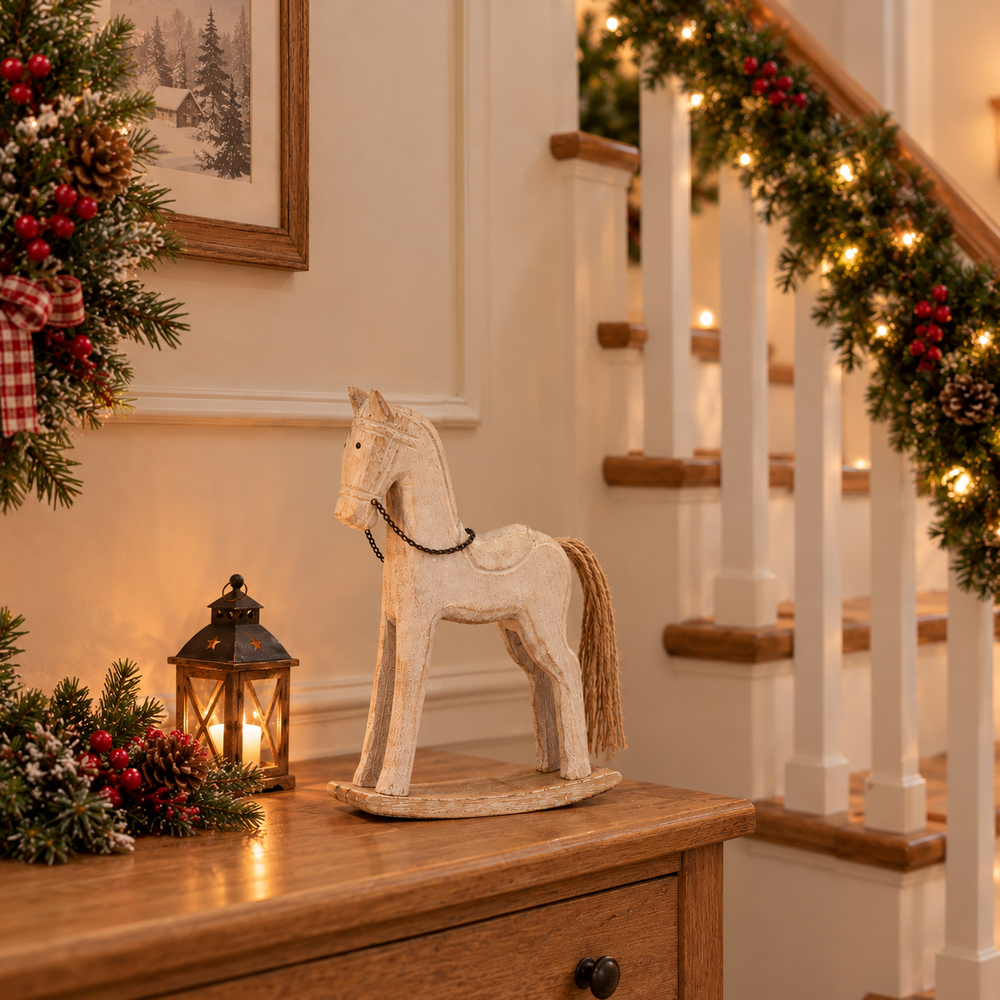 A rustic white wooden rocking horse with a natural brown mane and tail stands on a wooden console table in a warmly lit holiday home. The scene includes a lit lantern, green garlands with warm lights on the staircase railing, and festive wreaths.