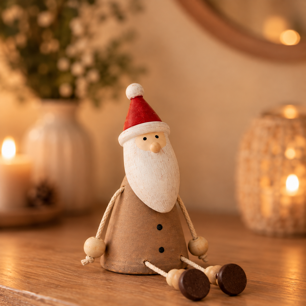 A small wooden Santa Claus figurine with a red hat, white beard, and brown body, posed on a wooden surface with warm blurred candles and decor in the background.