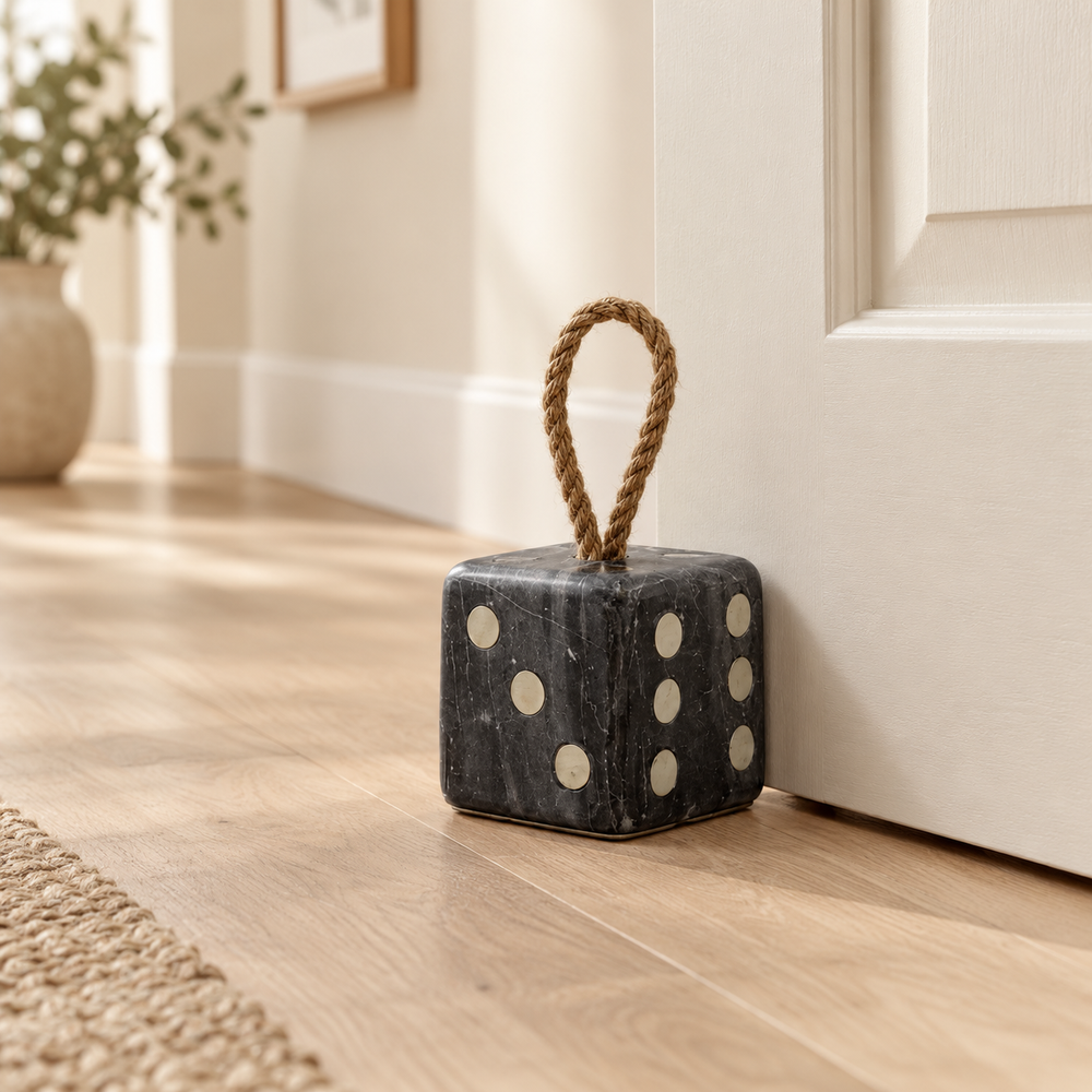 A dark gray marble dice-shaped door stopper with a natural jute rope handle, resting on a light wood floor next to a white door.