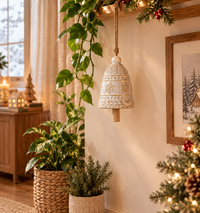 A decorative cream-colored ceramic Christmas bell ornament hangs from a string in a cozy, warmly lit living room decorated with potted plants, a Christmas tree, and holiday garlands.