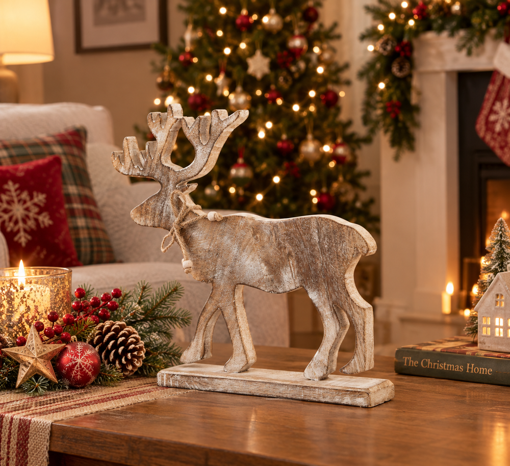 A rustic, weathered wood reindeer figurine with antlers stands on a matching rectangular base on a wooden table, set against a cozy Christmas living room background with a lit tree, fireplace, and holiday decorations.