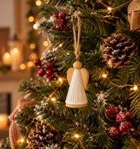 A ceramic angel Christmas ornament hangs from a decorated Christmas tree with warm string lights, pinecones, red berries, and snow-dusted green branches.