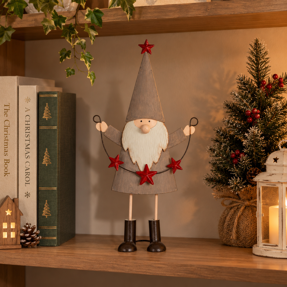 A Christmas gnome figurine with a gray hat and coat, white beard, and red stars, displayed on a wooden shelf next to books, a small tree, and a lantern.