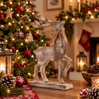 A rustic wooden reindeer figurine with antlers and a rope bow stands on a matching base on a wooden table, set against a warm, decorated Christmas tree and fireplace background.
