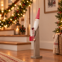 A tall wooden Christmas gnome figurine with a red and white striped hat, white beard, and red-and-white striped gift box, standing on a wooden floor in a decorated holiday home interior.