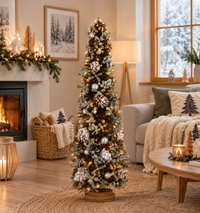 A decorated Christmas tree stands in a cozy living room with a fireplace, sofa, and warm ambient lighting. The tree is frosted with light snow-like branches and adorned with white and silver ornaments and pine cones.